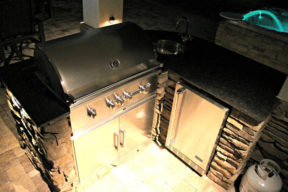 Outdoor kitchen with stainless steel grill, mini-fridge, sink, and stone facade, illuminated at night.