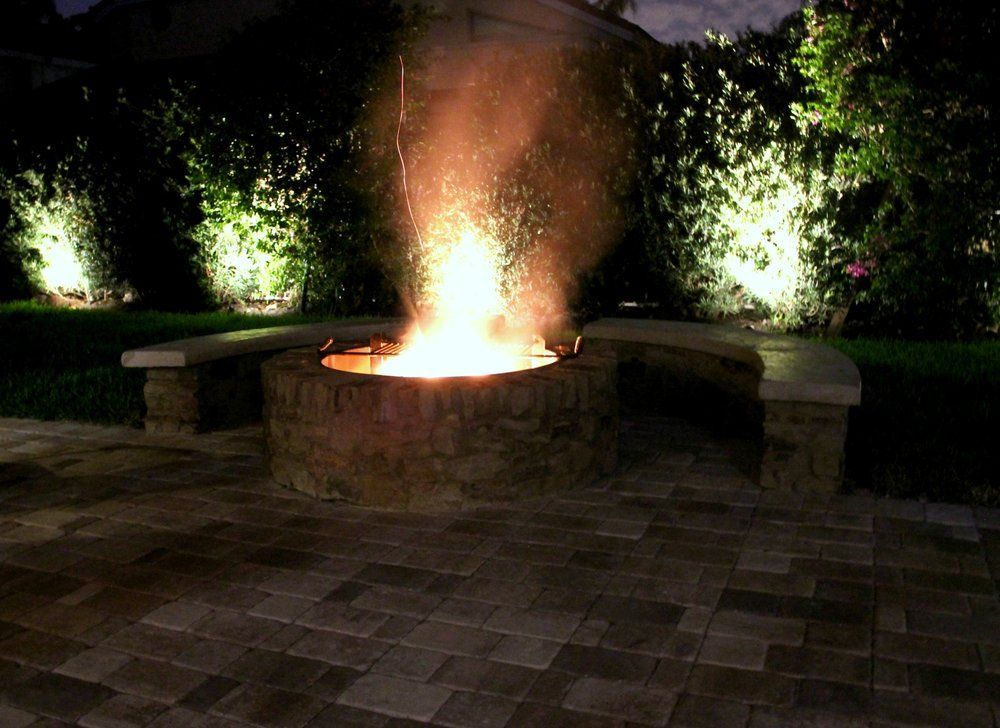 Fire pit burning brightly with stone benches, pavers, and illuminated greenery at night.