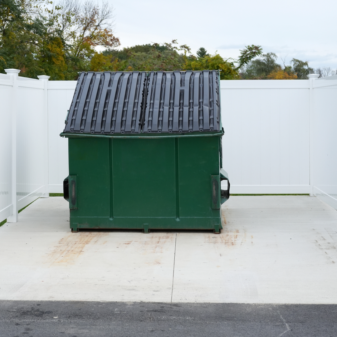 a green dumpster with a black roof sits in front of a white fence