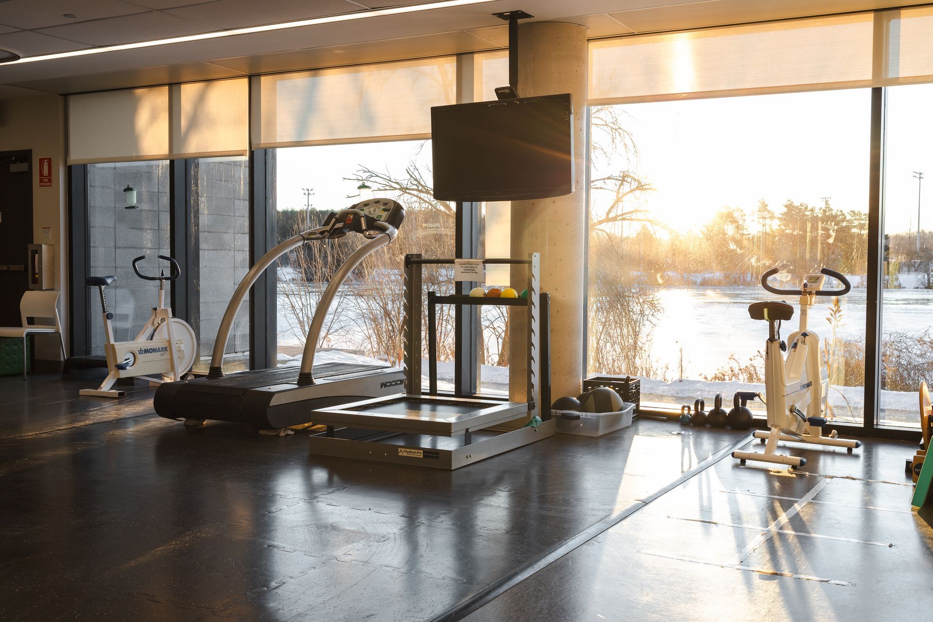 Gym interior with exercise equipment, a large window overlooking a snowy landscape, and a TV screen.