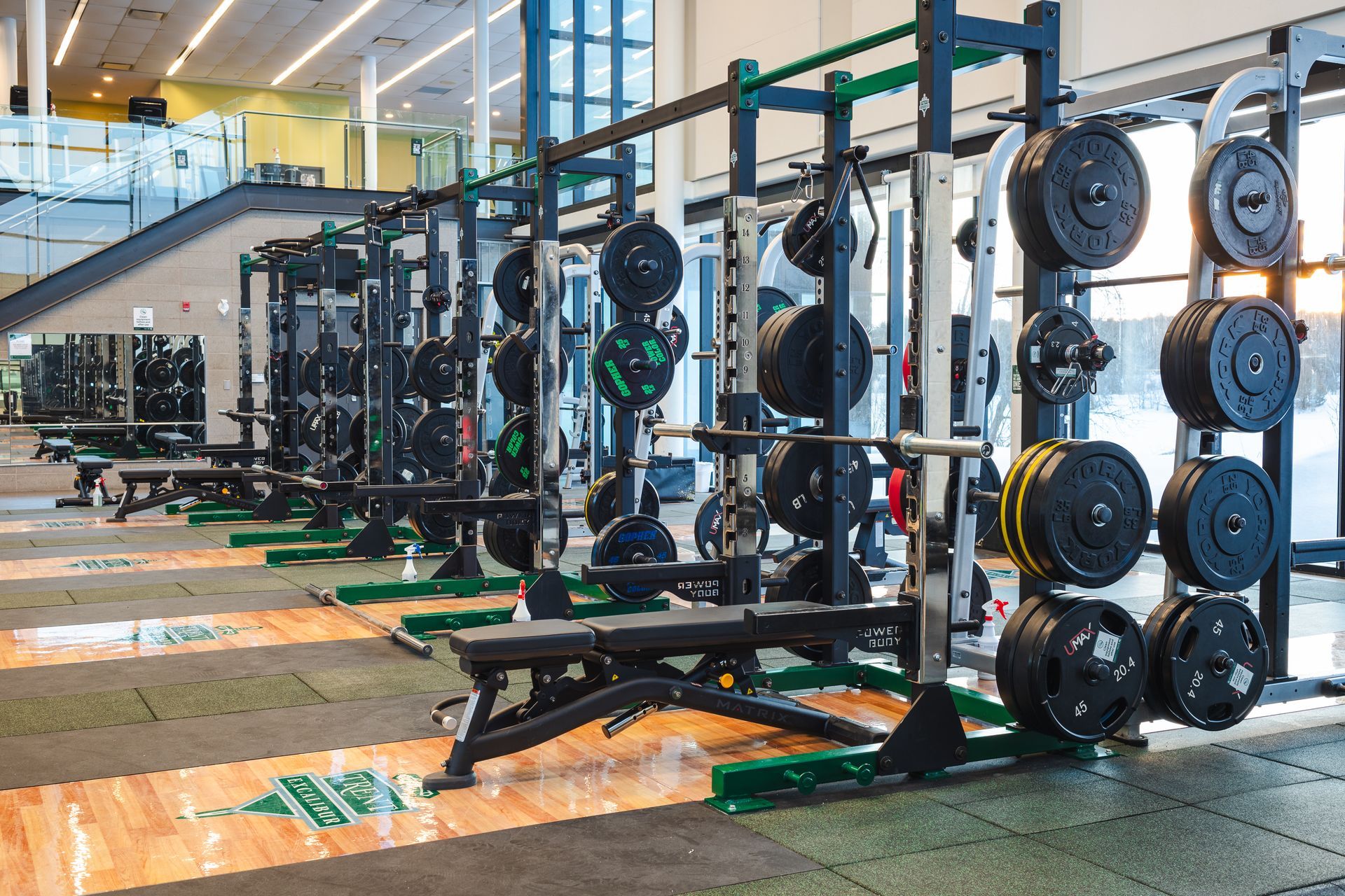 Weightlifting racks in a gym, loaded with weights. Benches, green and black mats, bright windows.