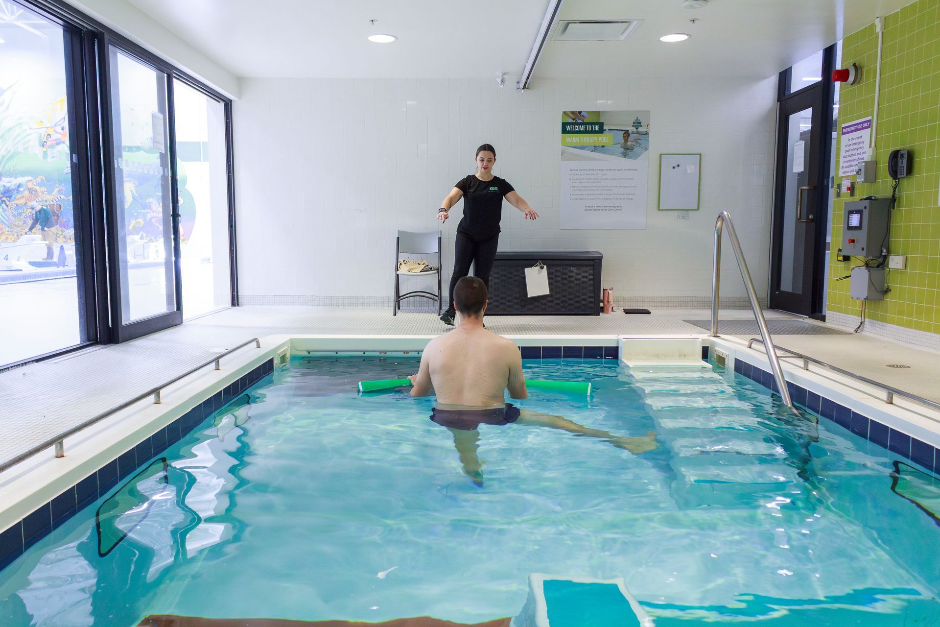 A man in a pool doing leg lifts under the supervision of a trainer in a rehabilitation center.