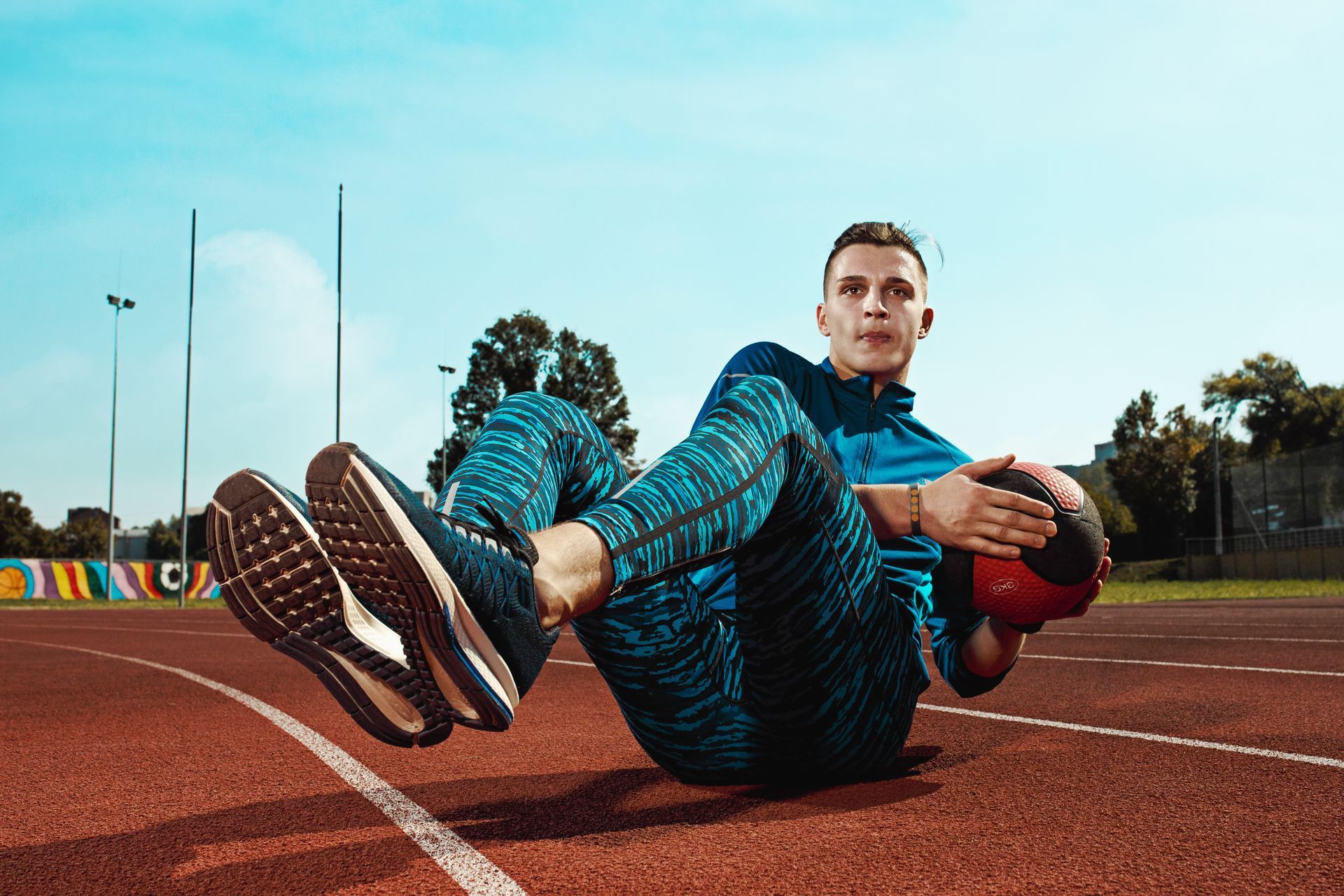Man on a track doing a Russian twist with a medicine ball, blue sky background.