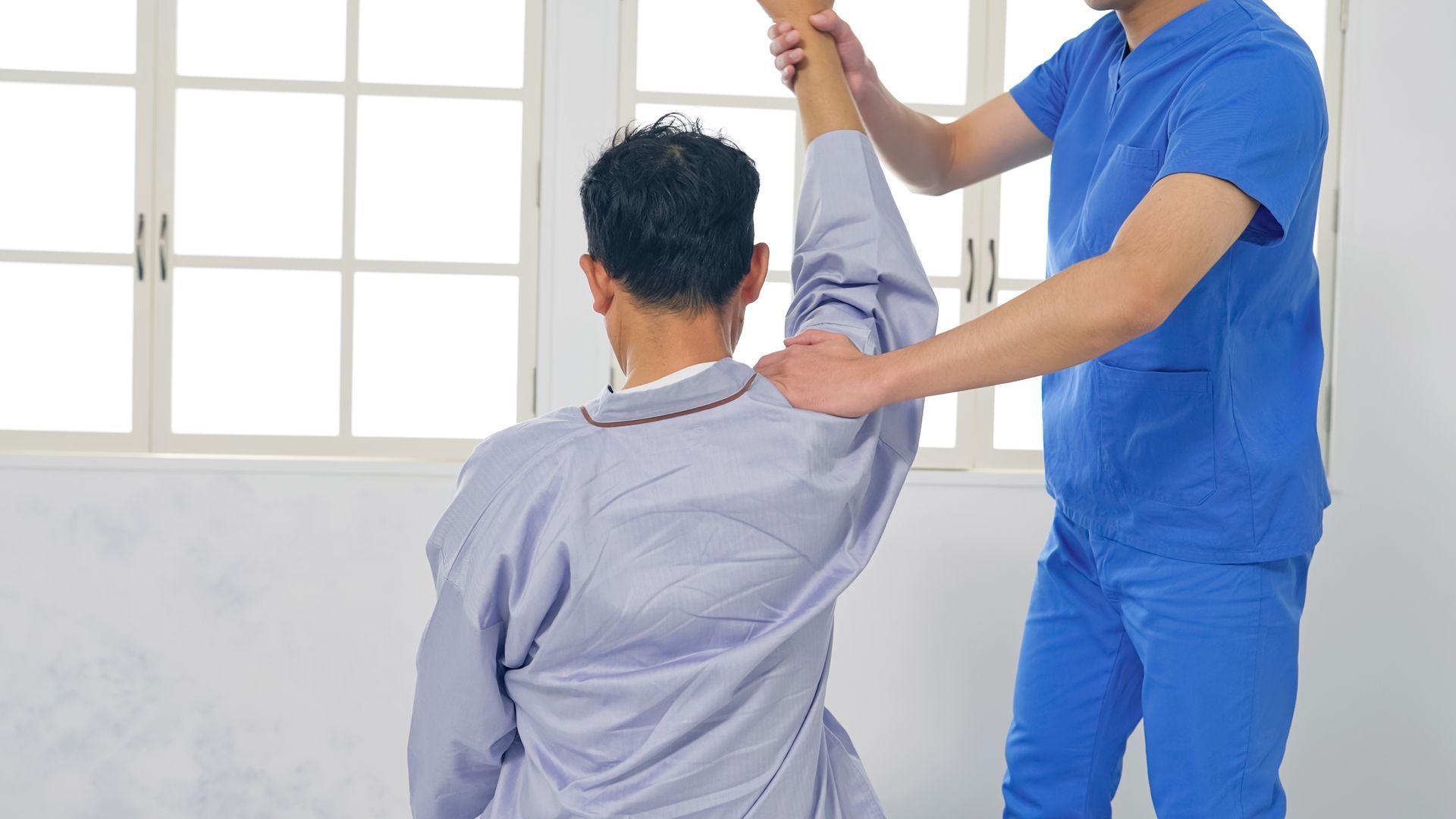 Therapist in blue scrubs stretches a patient's arm in front of a window.