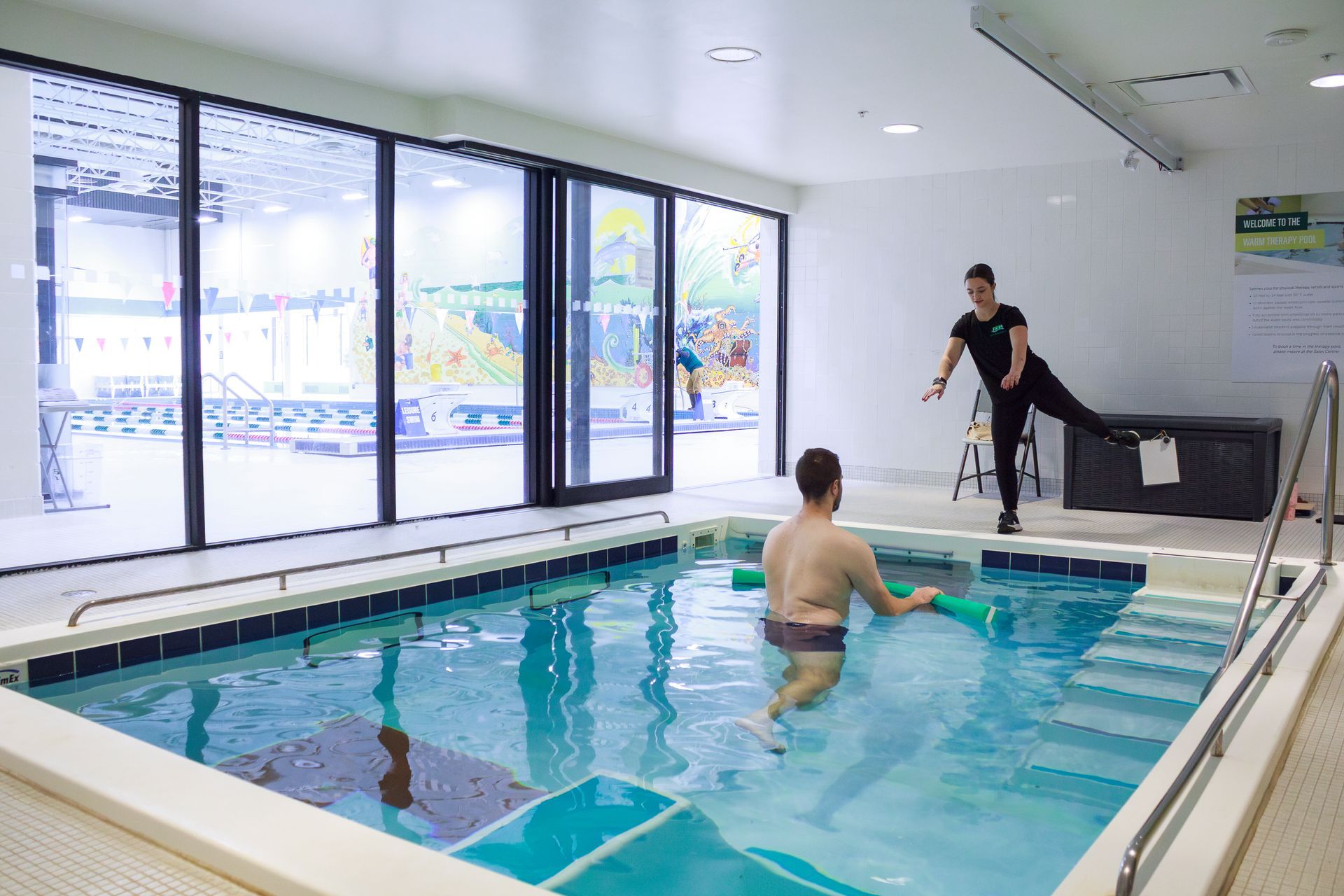Man in therapy pool with trainer guiding exercise. White room, large windows.