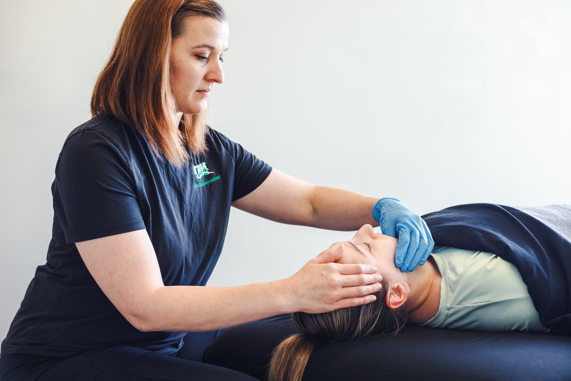 Woman giving a massage to another person's head, indoors. Both are caucasian. The massage giver wears gloves and a dark shirt.