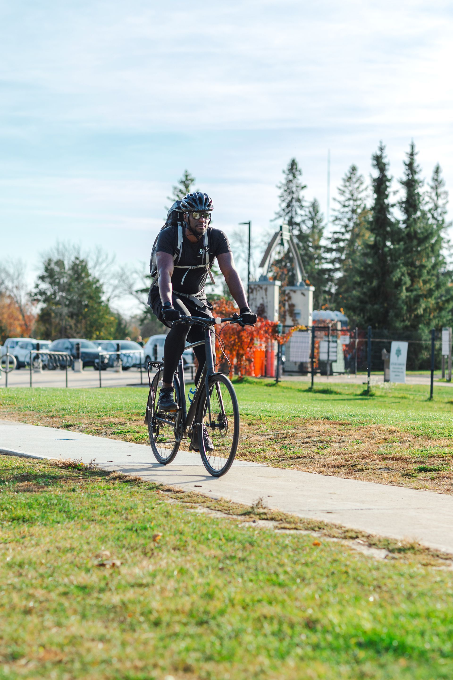 Man on bike riding on a paved path. He wears a helmet and backpack in a park on a sunny day.