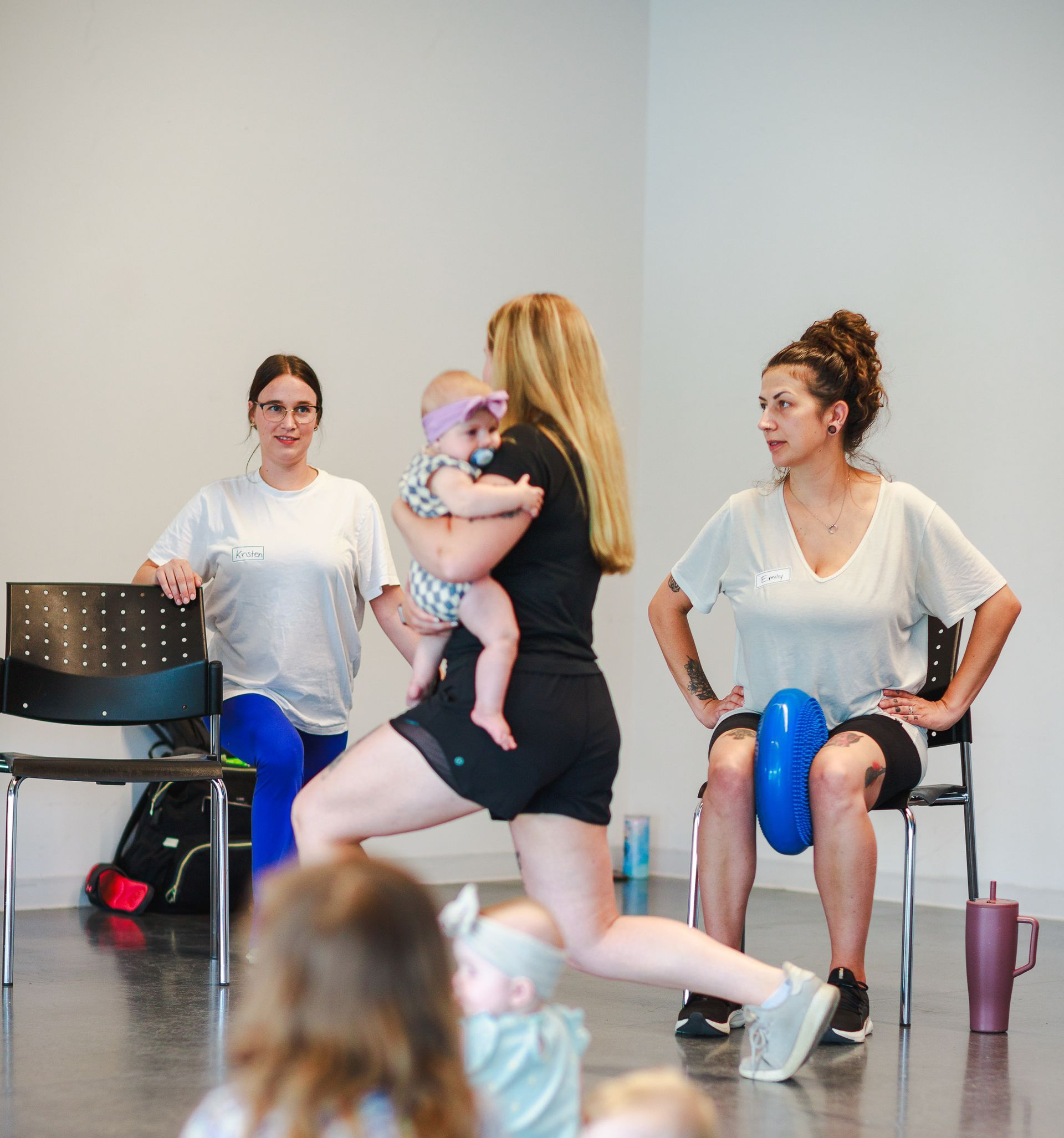 Women exercising with babies in a brightly lit room. One woman does lunges, another sits on a chair with a ball between her legs.