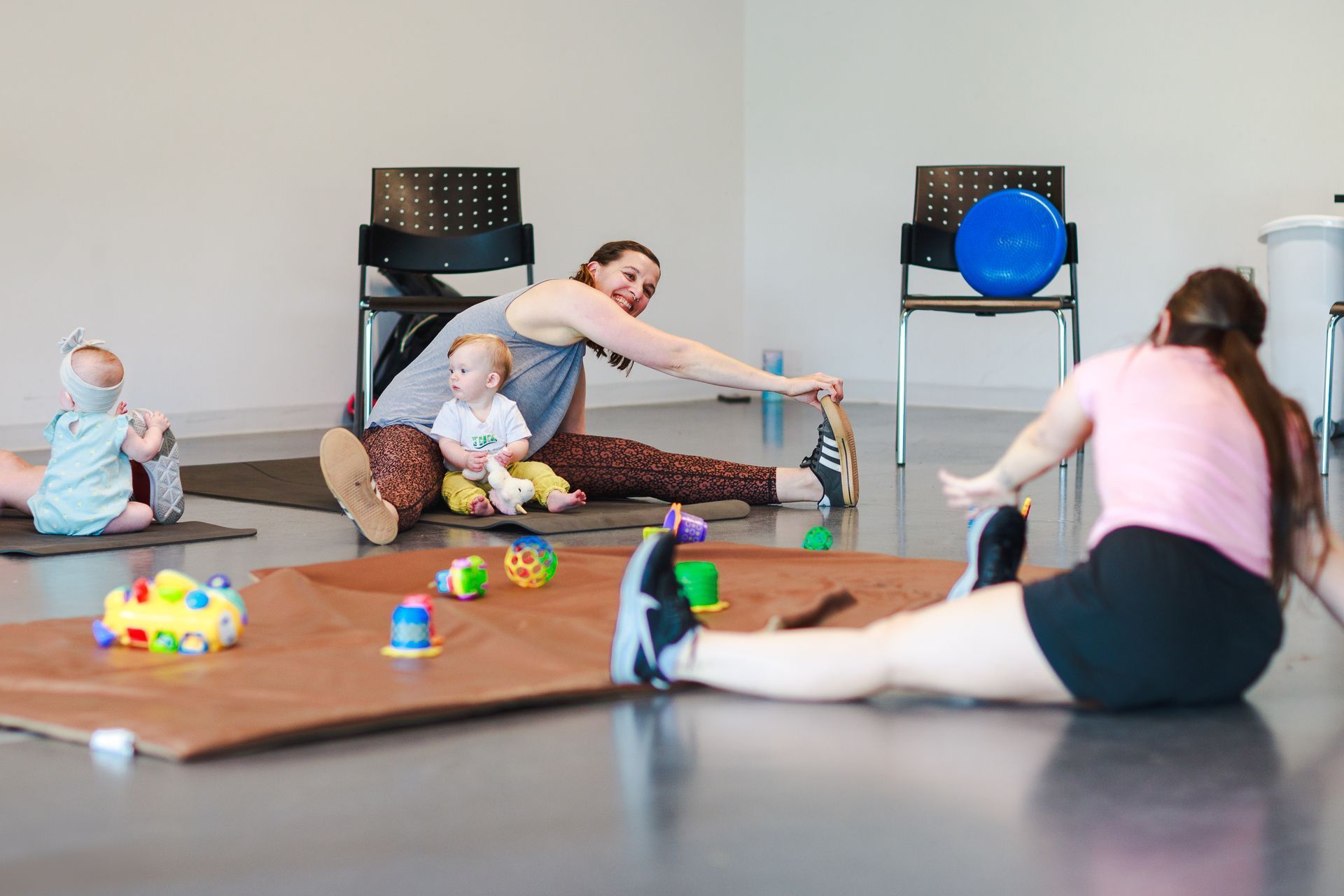 Women stretching with babies at a fitness class, indoors. Floor mats, toys, and a blue exercise ball are present.