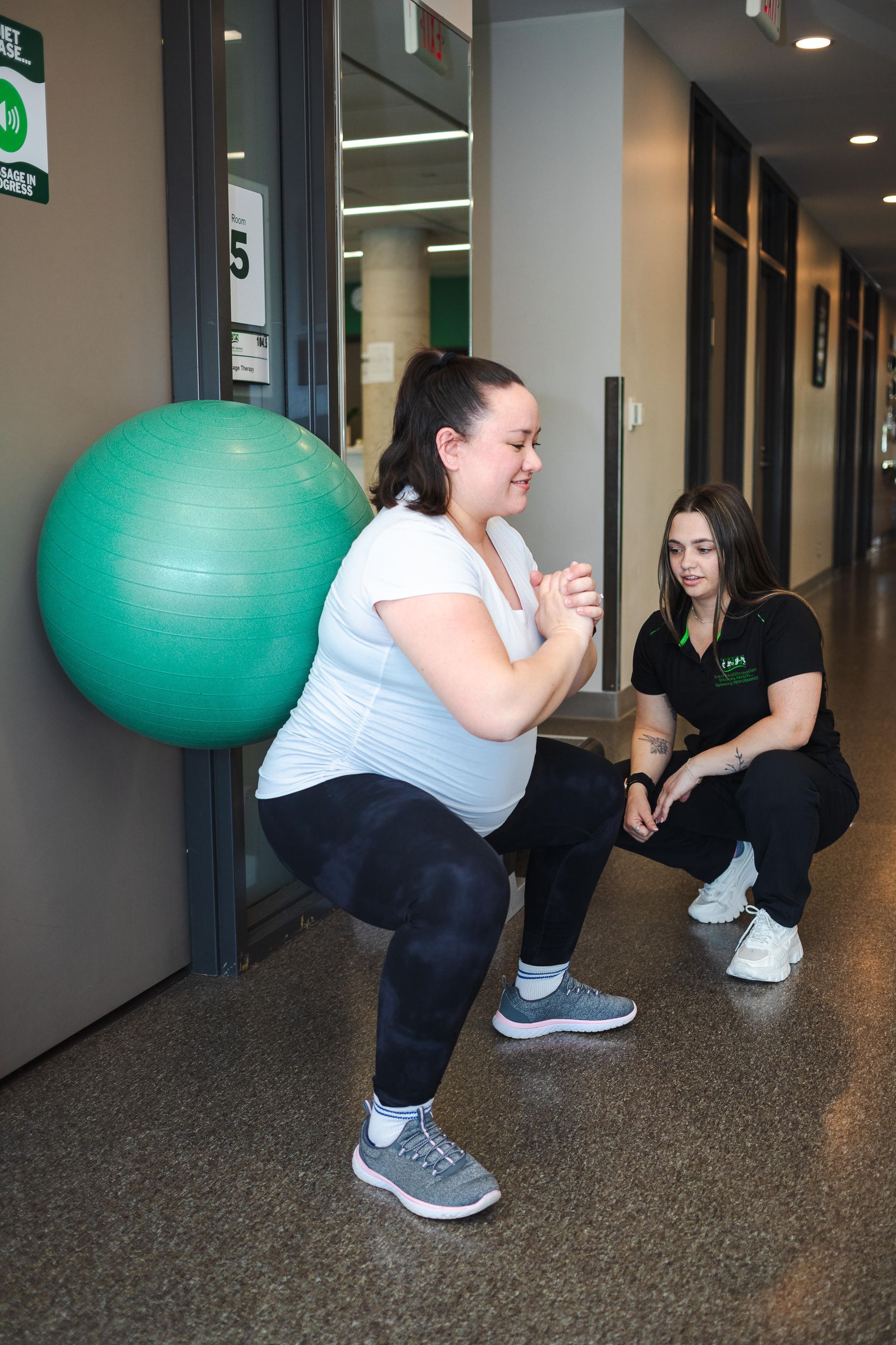 Pregnant woman squats against ball, guided by trainer in a hallway.