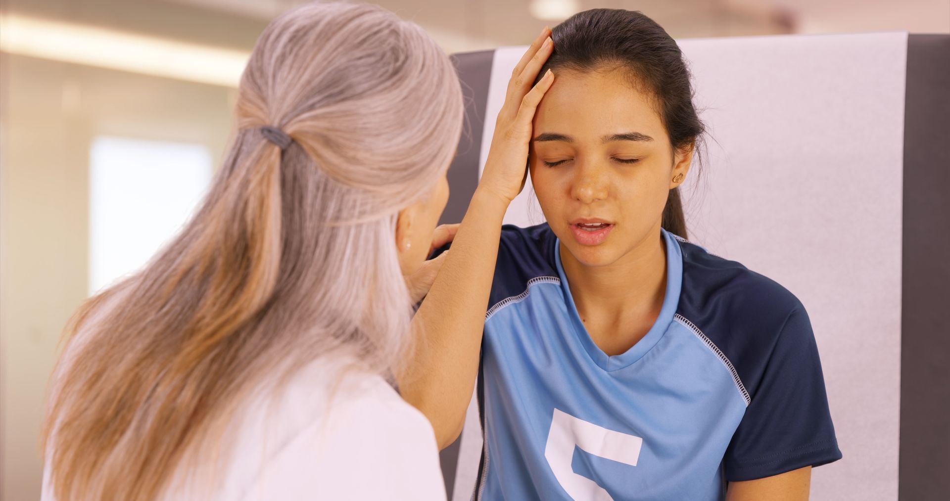 Woman in sports uniform being examined by a doctor, who is touching her forehead; indoor setting.