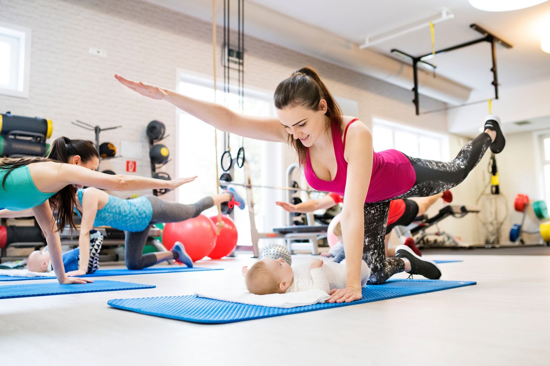 Women in a gym do fitness with a baby, one woman balances an arm and leg.