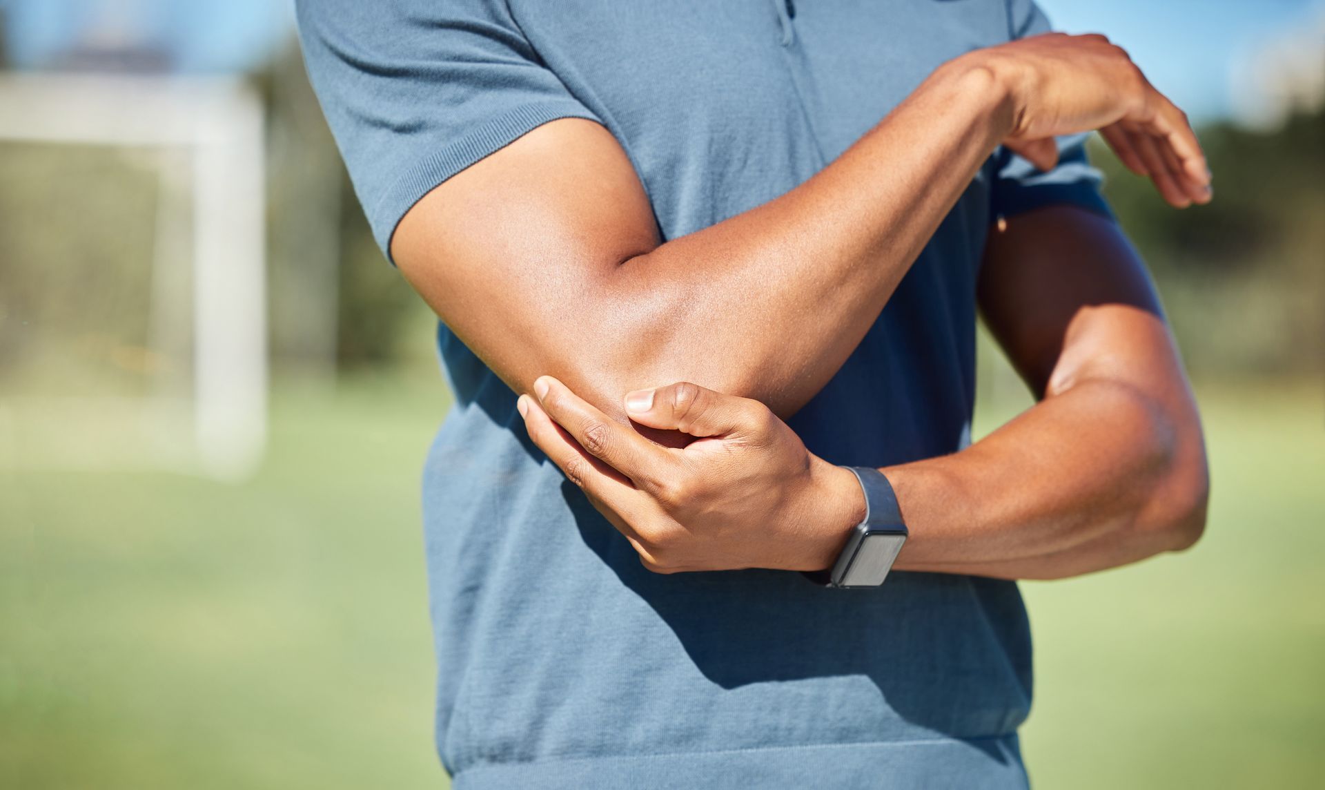 Man holding his elbow, likely in pain, outdoors. Wearing a blue shirt.