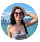 Mulher de óculos escuros e chapéu sorri, com o braço levantado, perto da água, com prédios ao longe.