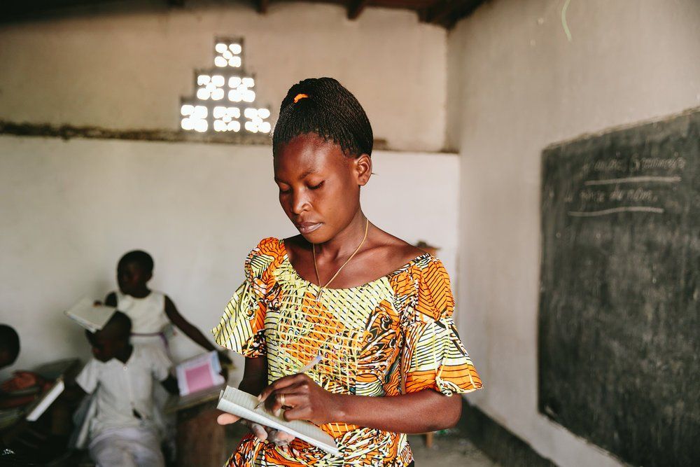 Girl in colorful dress writing in notebook in a classroom; other students seated in background.