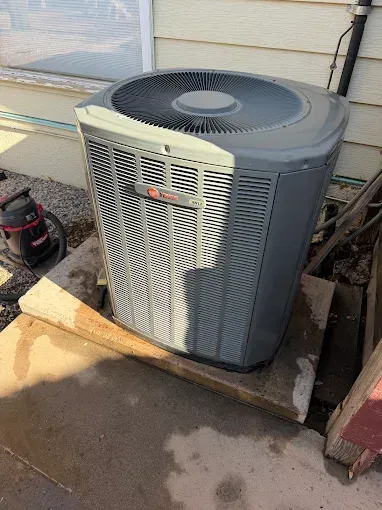 Outdoor gray HVAC condenser unit beside a house, with a shadow and wet pavement nearby