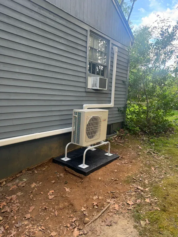 Exterior air conditioning unit on a raised platform next to a gray house, with a window unit above.