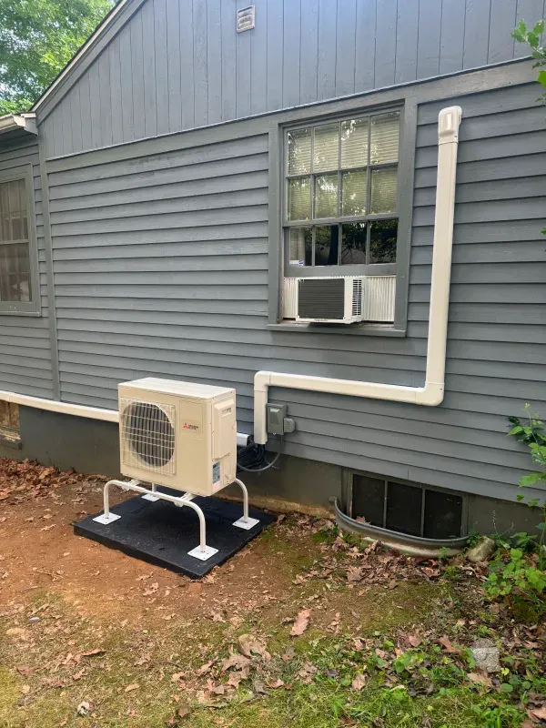 Outdoor HVAC unit on a platform next to a gray house with a window AC unit and vent pipes.