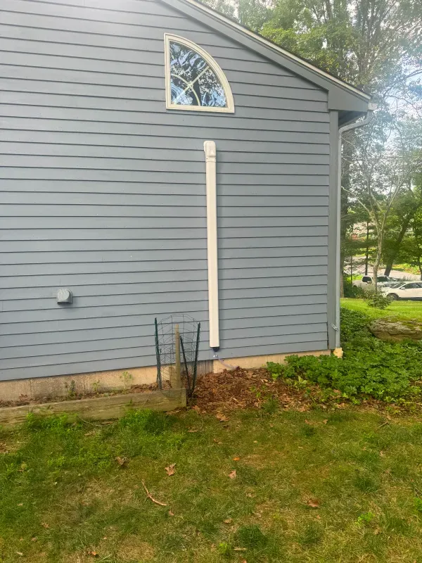 Blue siding of a house with a white downspout and arched window. Green grass and shrubs in foreground.