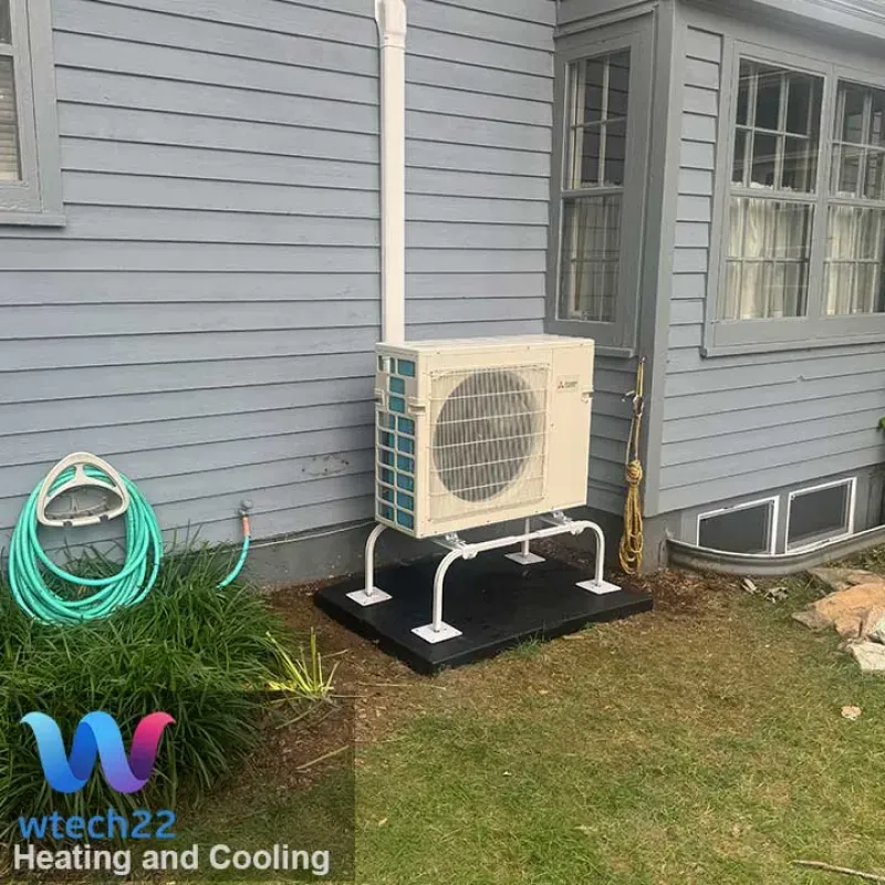 An air conditioning unit on a raised platform next to a blue house with a window, grass and a hose.