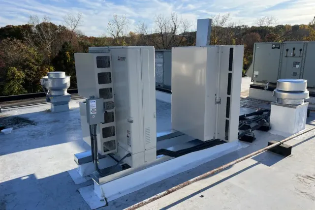 White HVAC units on a flat roof with vents and trees in the background under a blue sky.