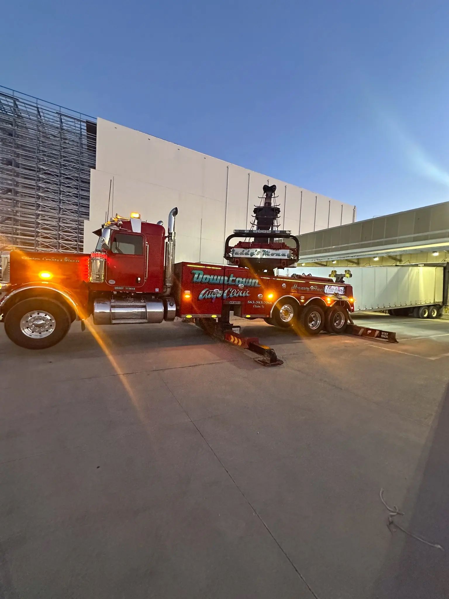 Red semi-truck with a boom lifting a cargo container outside a white warehouse at dusk | Downtown Auto Clinic