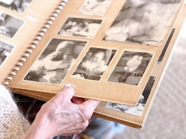 An elderly woman is holding a photo album full of black and white photos.