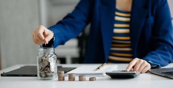 A woman is putting coins into a jar while using a calculator.