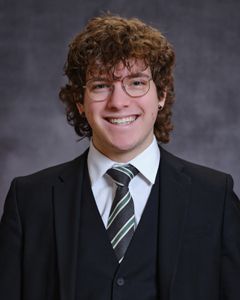 Man with curly brown hair, glasses, suit, and tie smiling for a portrait.