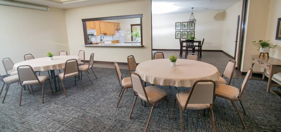 A room with tables and chairs in it and a kitchen in the background.