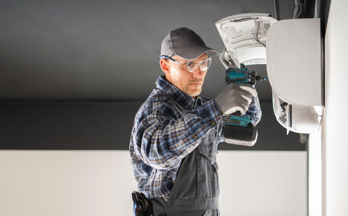 Man in workwear uses a drill on a white appliance near a black ceiling.