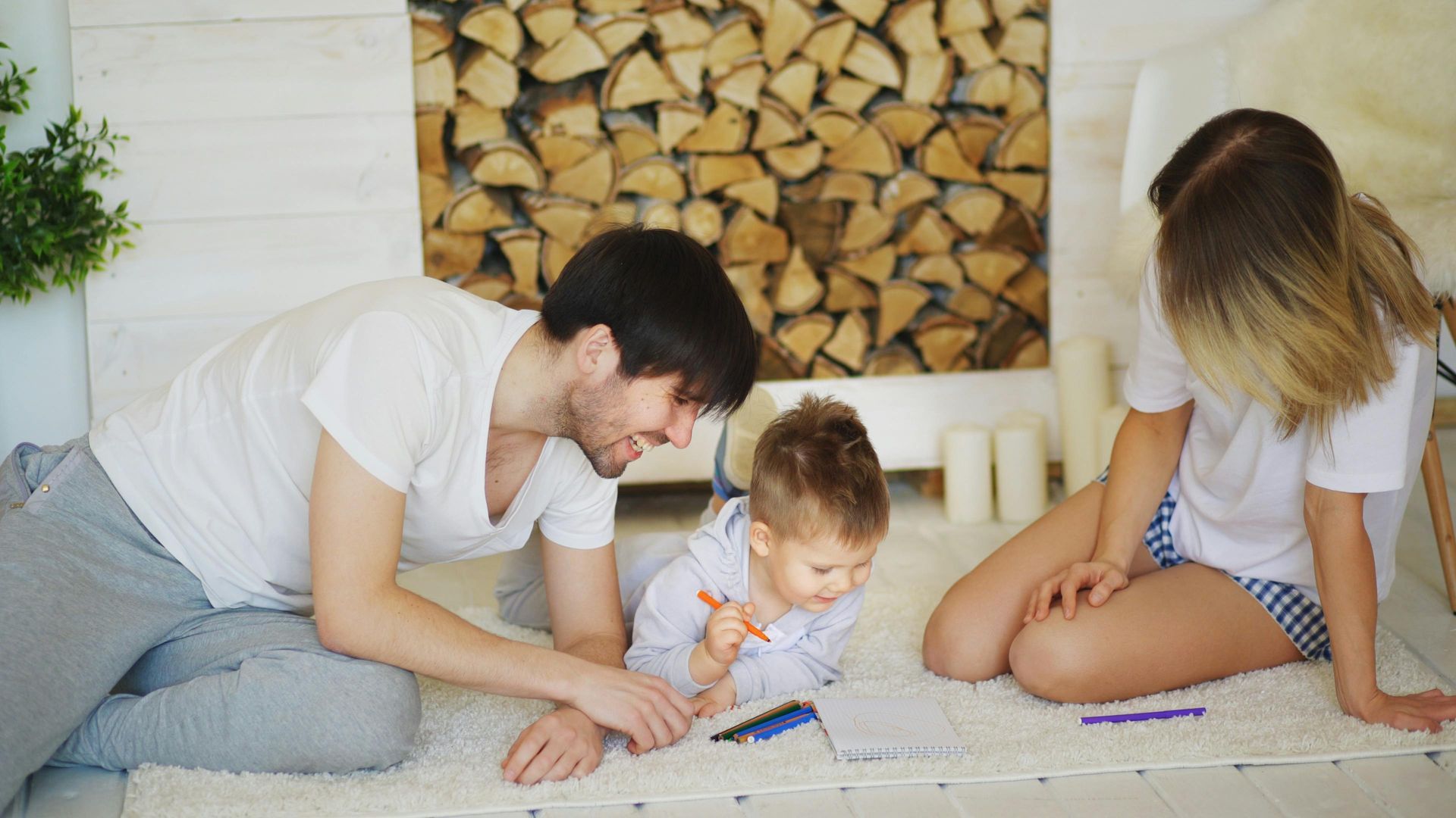 Family drawing on a white rug: Man and woman in casual clothes watch a child coloring with crayons, fireplace in background.