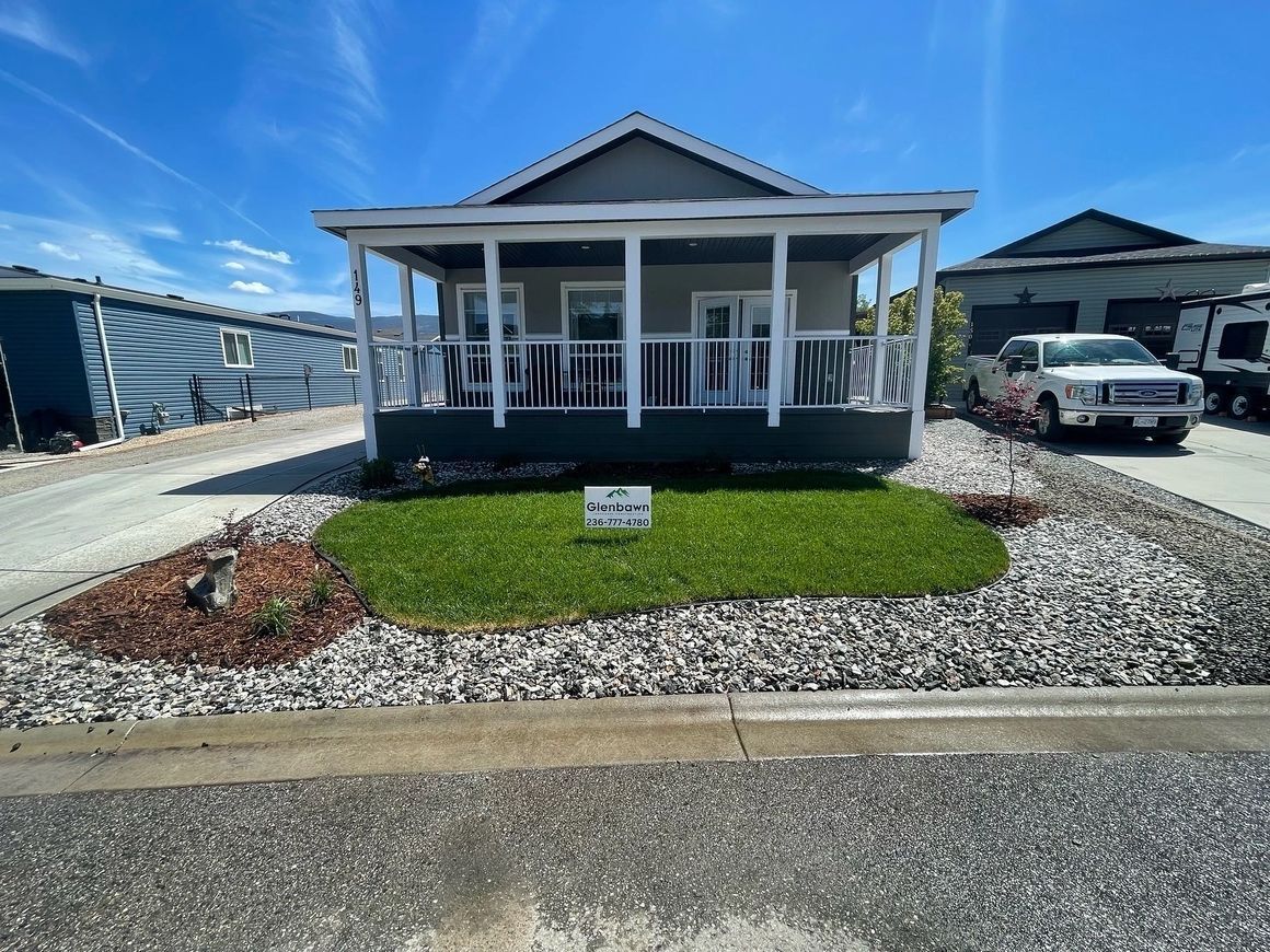 Small gray house with white porch and green lawn, surrounded by gravel, under a blue sky.
