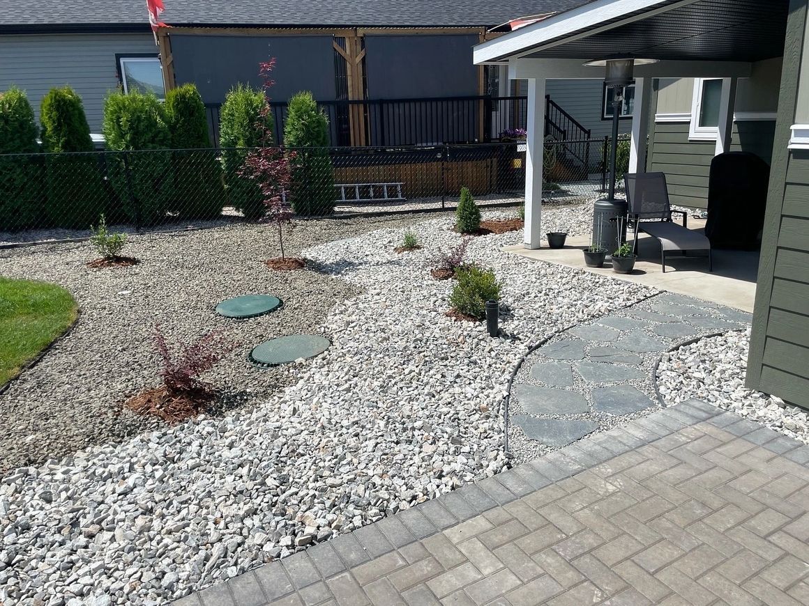 Landscaped yard with gravel, stepping stones, and small trees next to a patio and green building.
