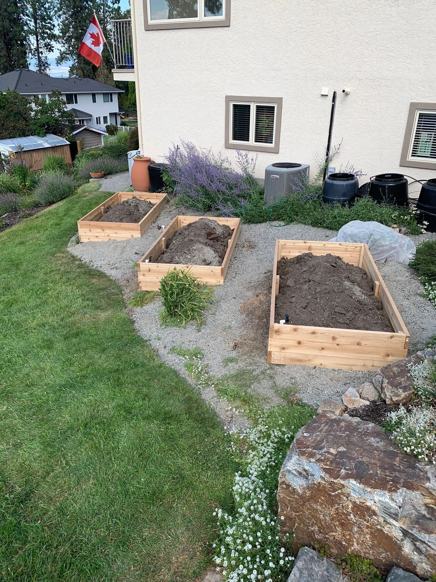 Three wooden raised garden beds filled with soil, set on gravel near a house with a Canadian flag.