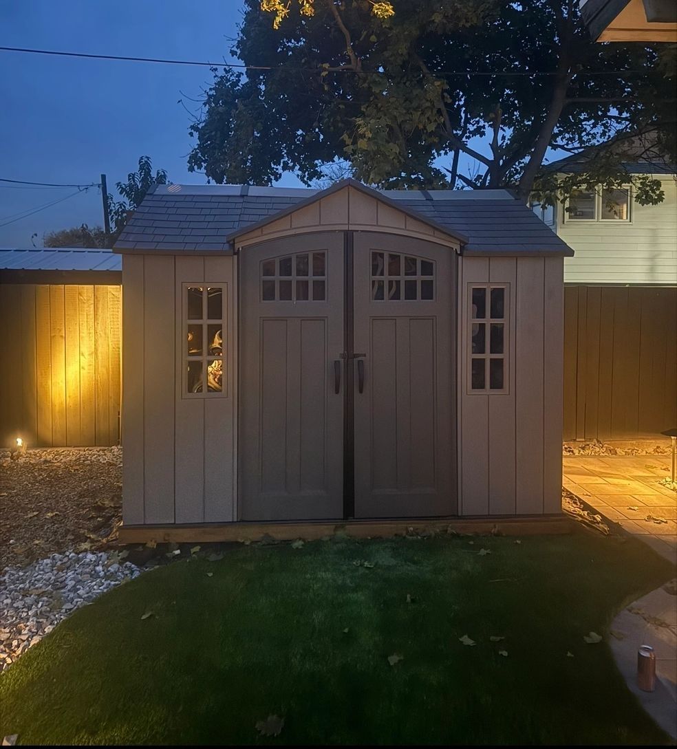Tan shed with gray roof and doors, two small windows, on grass next to a gravel path, nighttime.