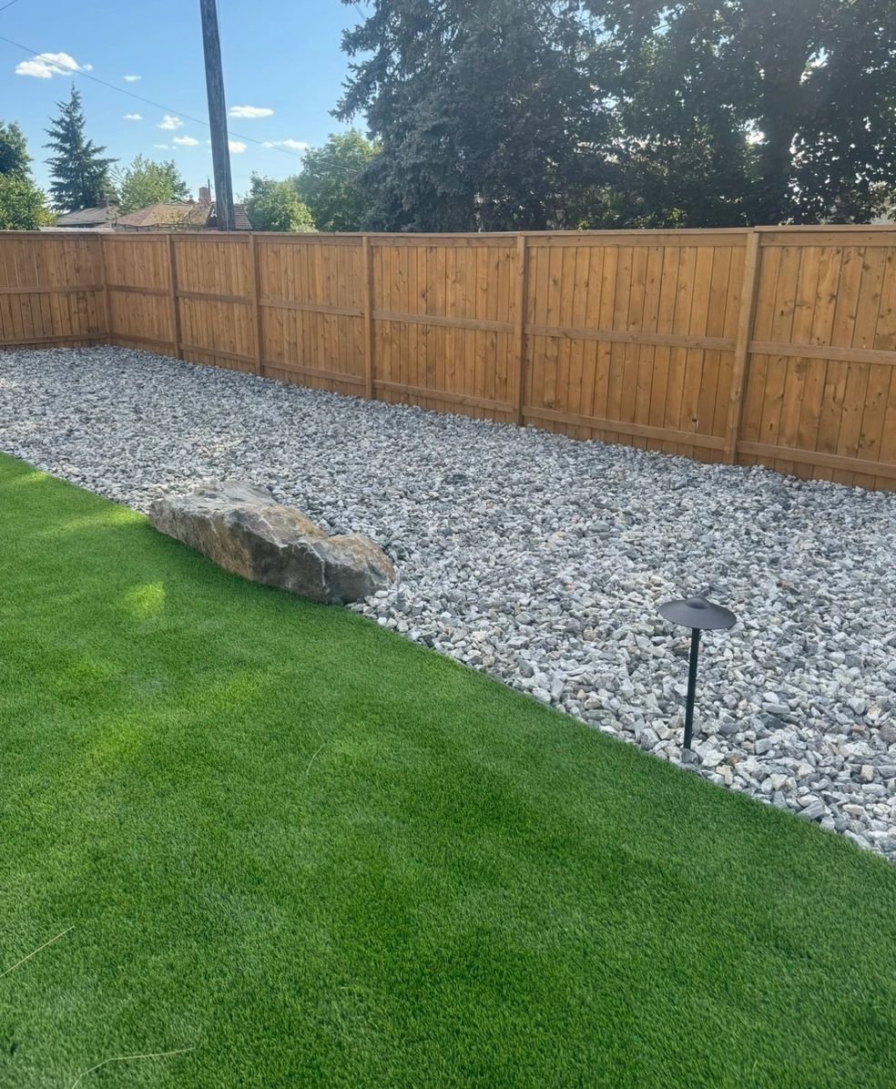 Green lawn and gravel area next to a wooden fence. A large rock sits on the lawn.