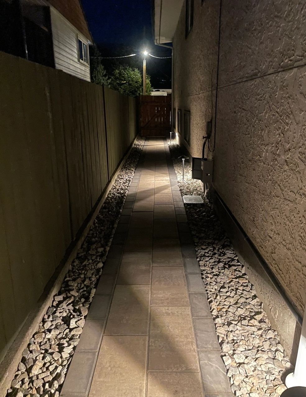 Narrow pathway between two walls, lit at night. Stone walkway with gravel borders leads to a gate.