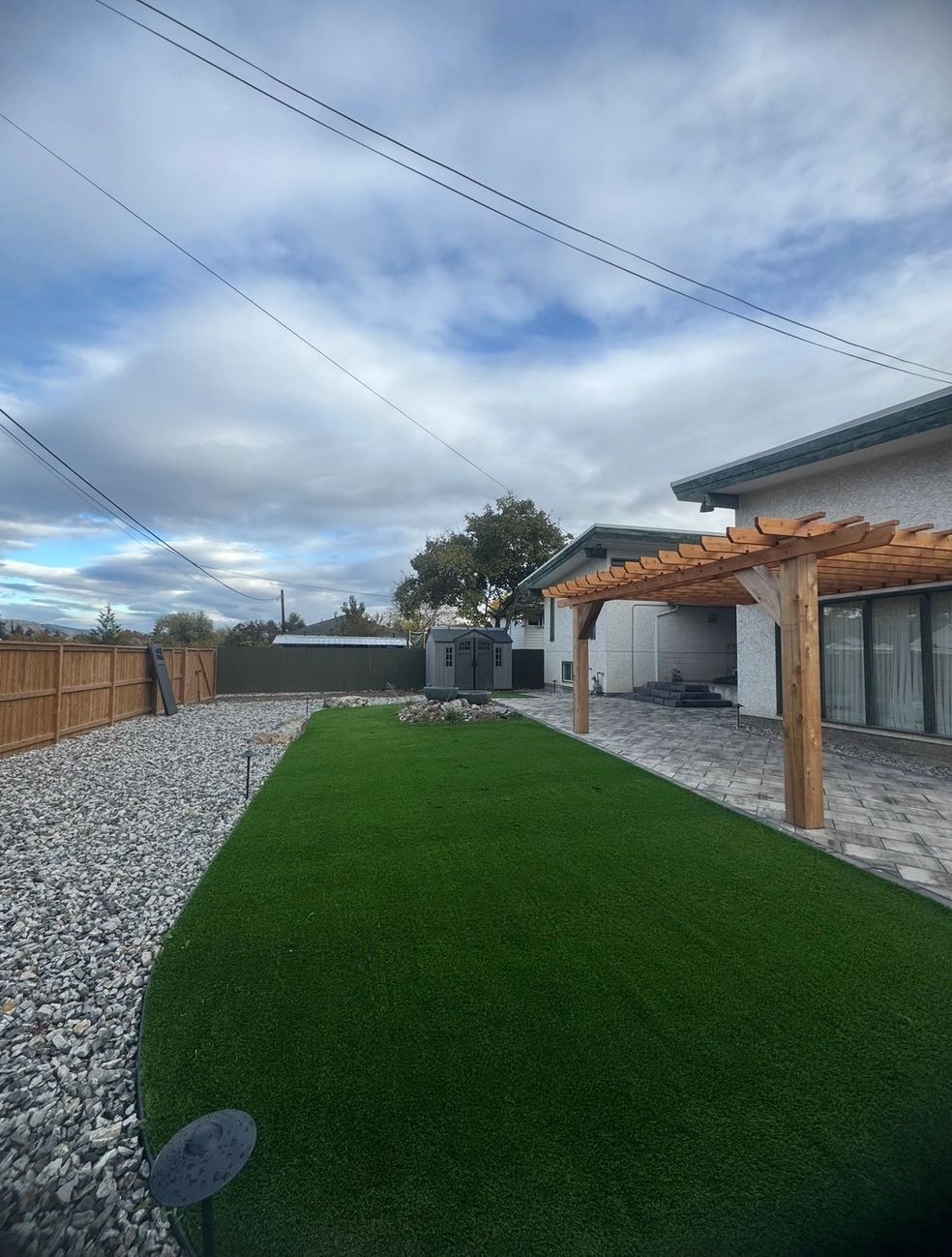 Backyard with artificial grass, gravel, wooden pergola, and cloudy sky.