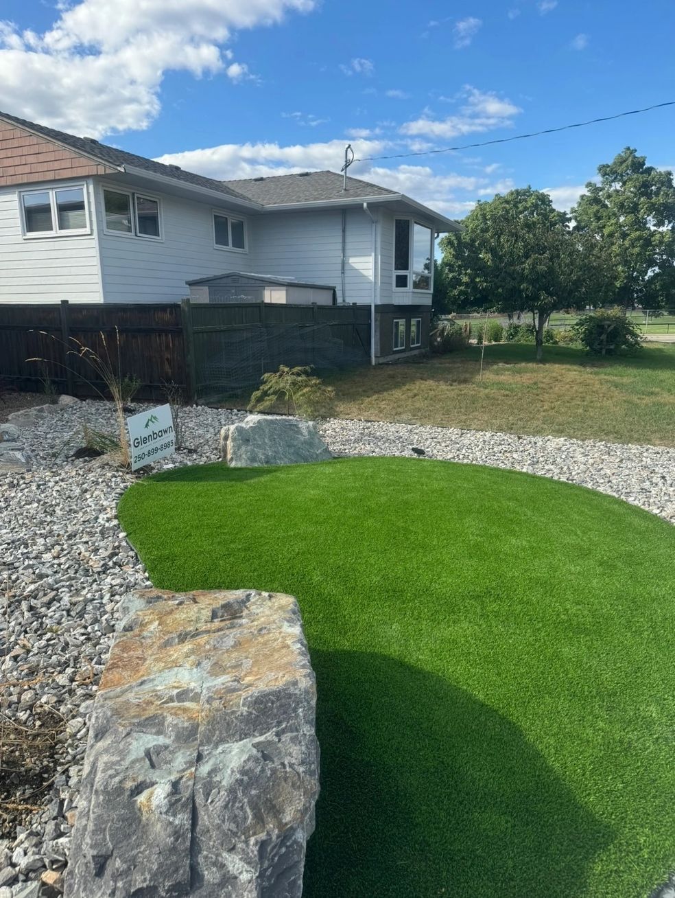 A green turf lawn bordered by rocks and gravel in front of a white house with a brown roof under a blue sky.