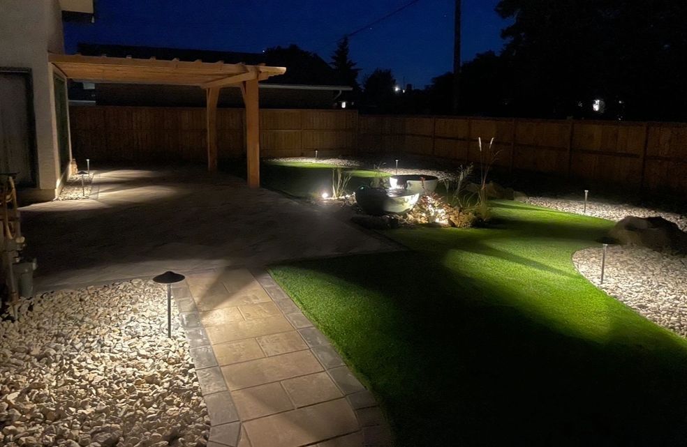 Backyard at night with landscape lighting illuminating a patio, lawn, and pergola.