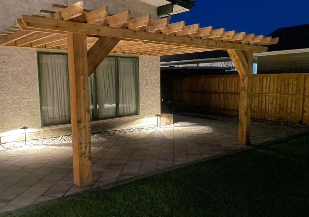 Wooden pergola over a stone patio, with illuminated spotlights and wooden fence in the background.