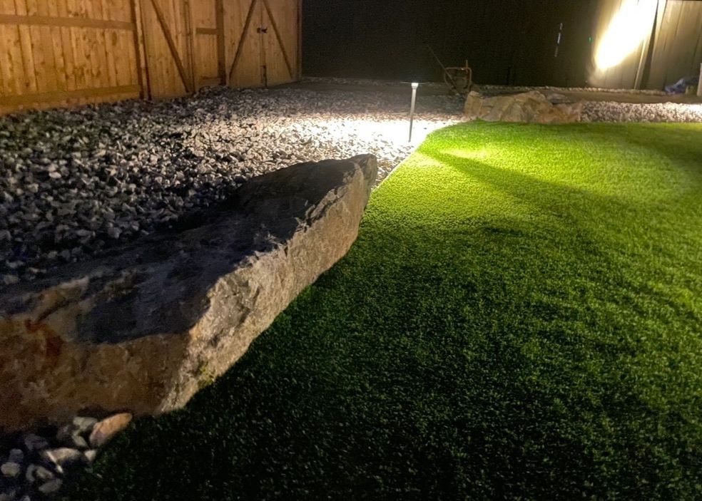 Large rock on gravel next to illuminated artificial turf. Backyard at night with wooden fence.