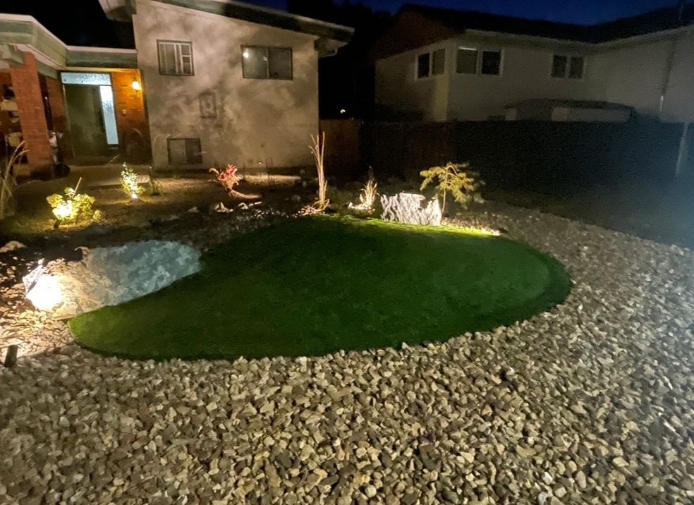 Night view of a yard with artificial grass, rocks, and illuminated landscaping around a home.