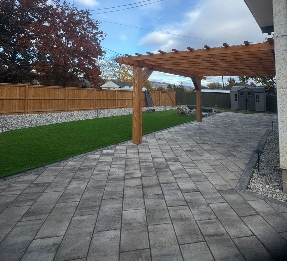 Patio with gray pavers, pergola, green lawn, wooden fence, and shed.