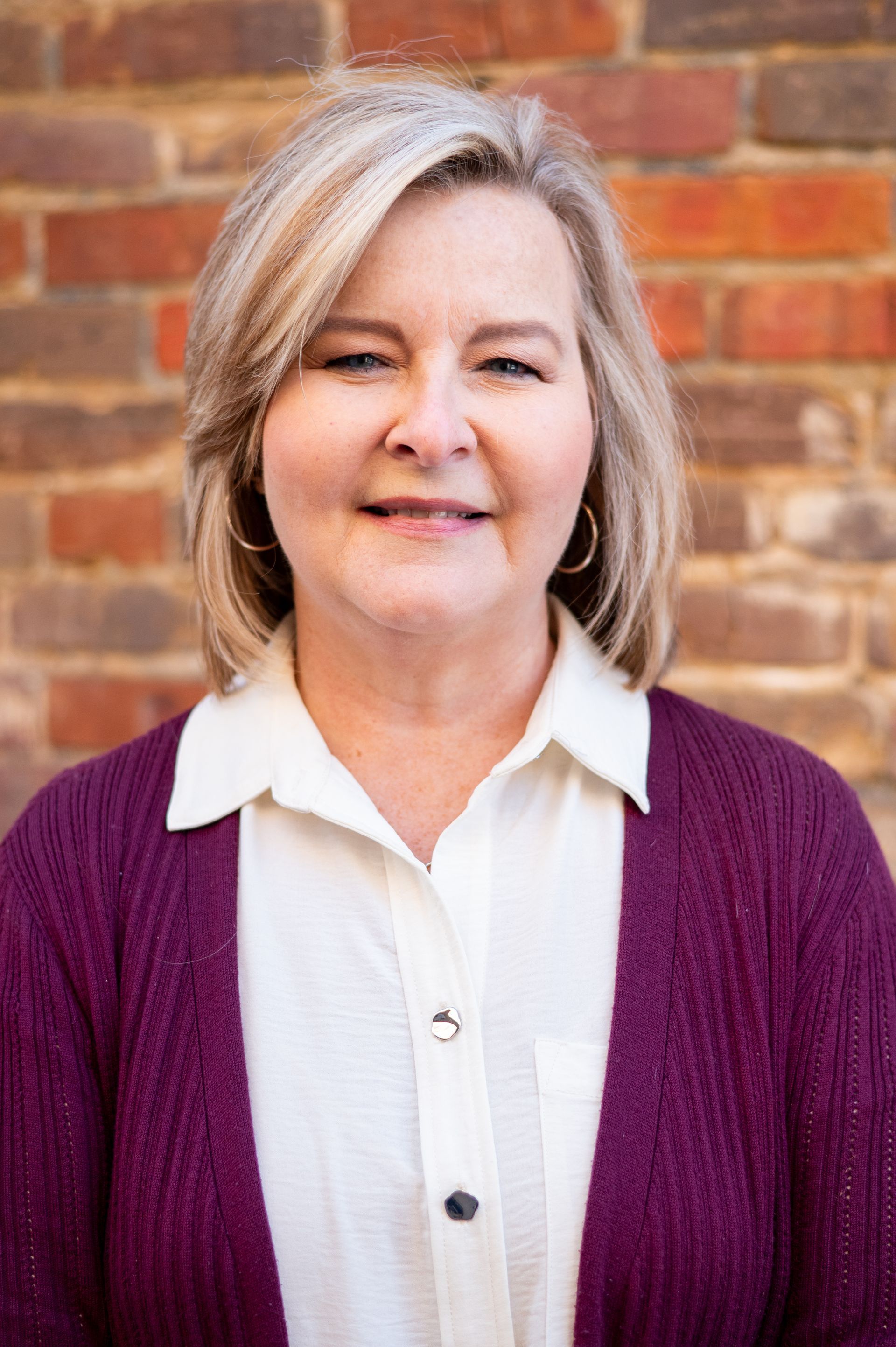 Woman with blonde hair wearing a purple cardigan and white shirt smiles in front of a brick wall.