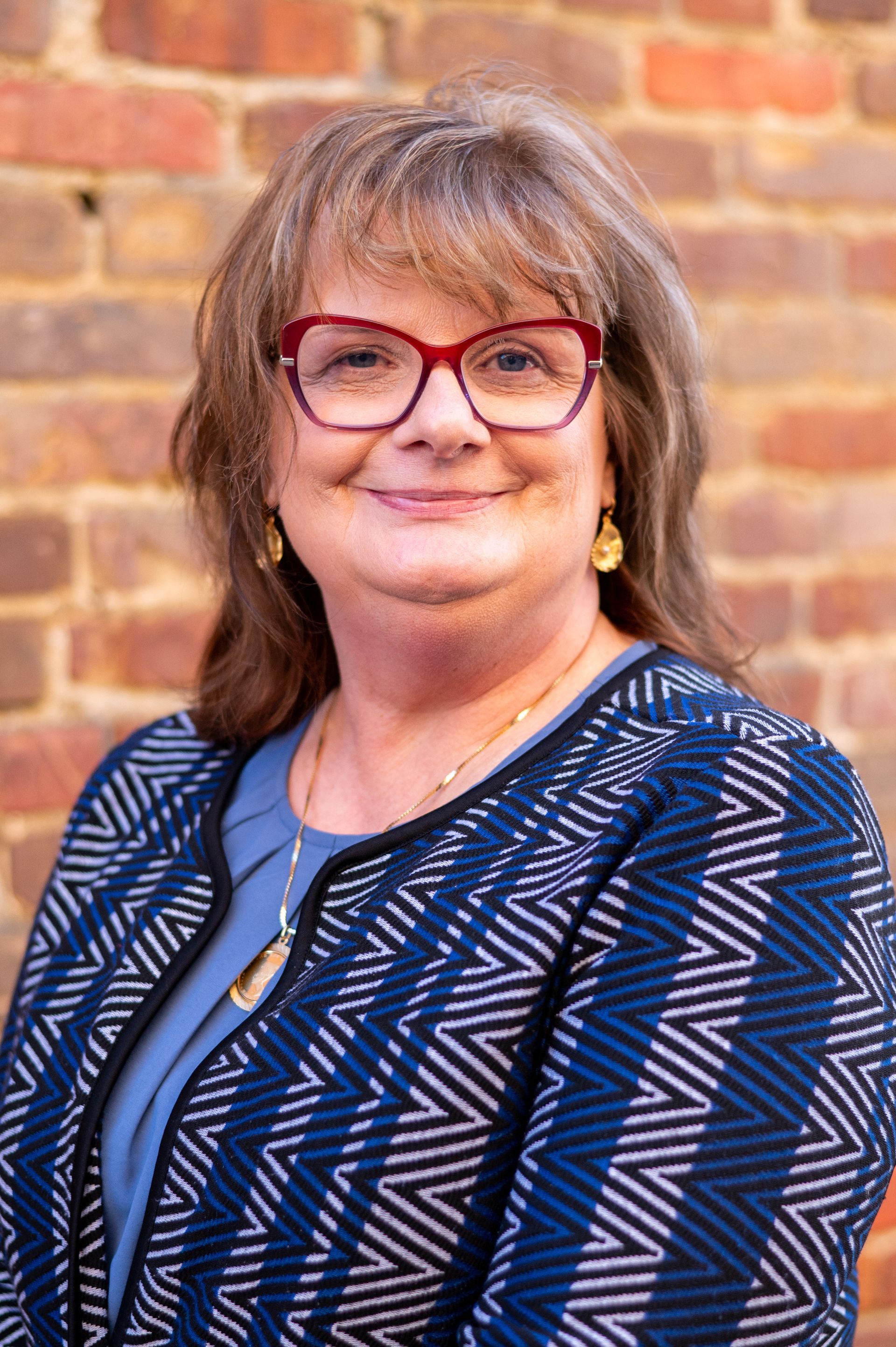 Woman with red glasses smiles, stands in front of a brick wall, wearing a patterned blazer.