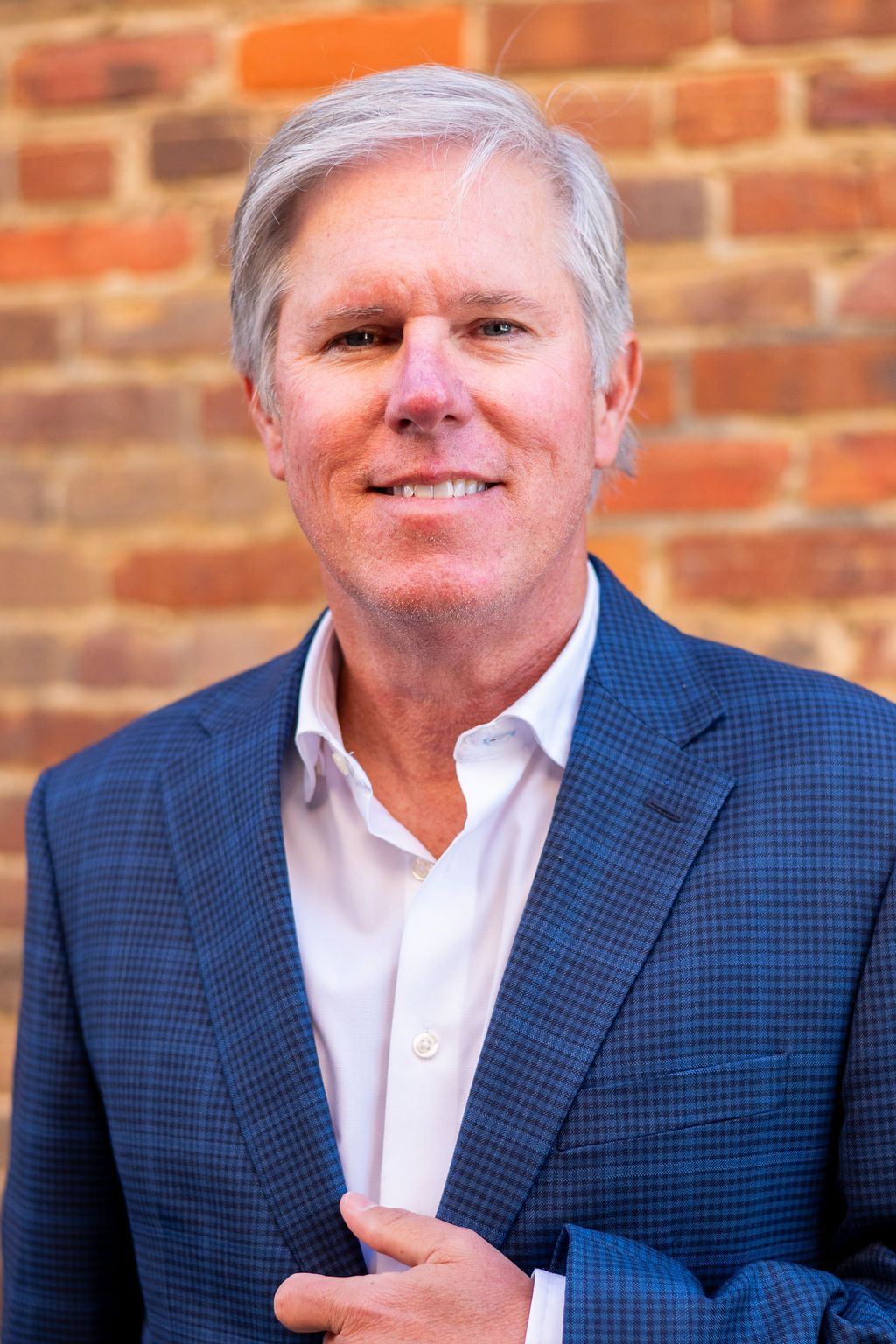 Man in blue blazer and white shirt smiles in front of a brick wall.