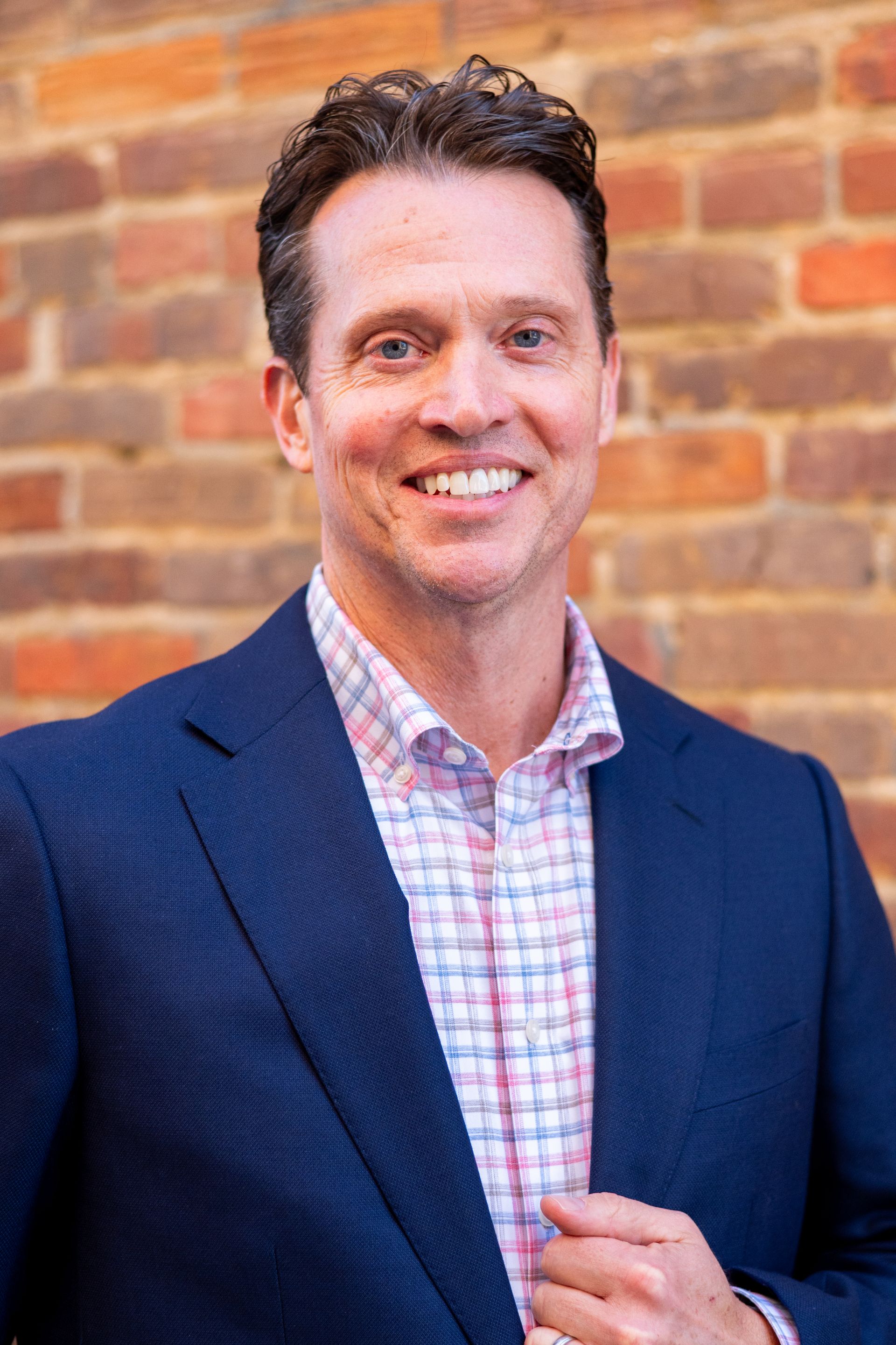 Man in navy blazer and patterned shirt smiling in front of a brick wall.