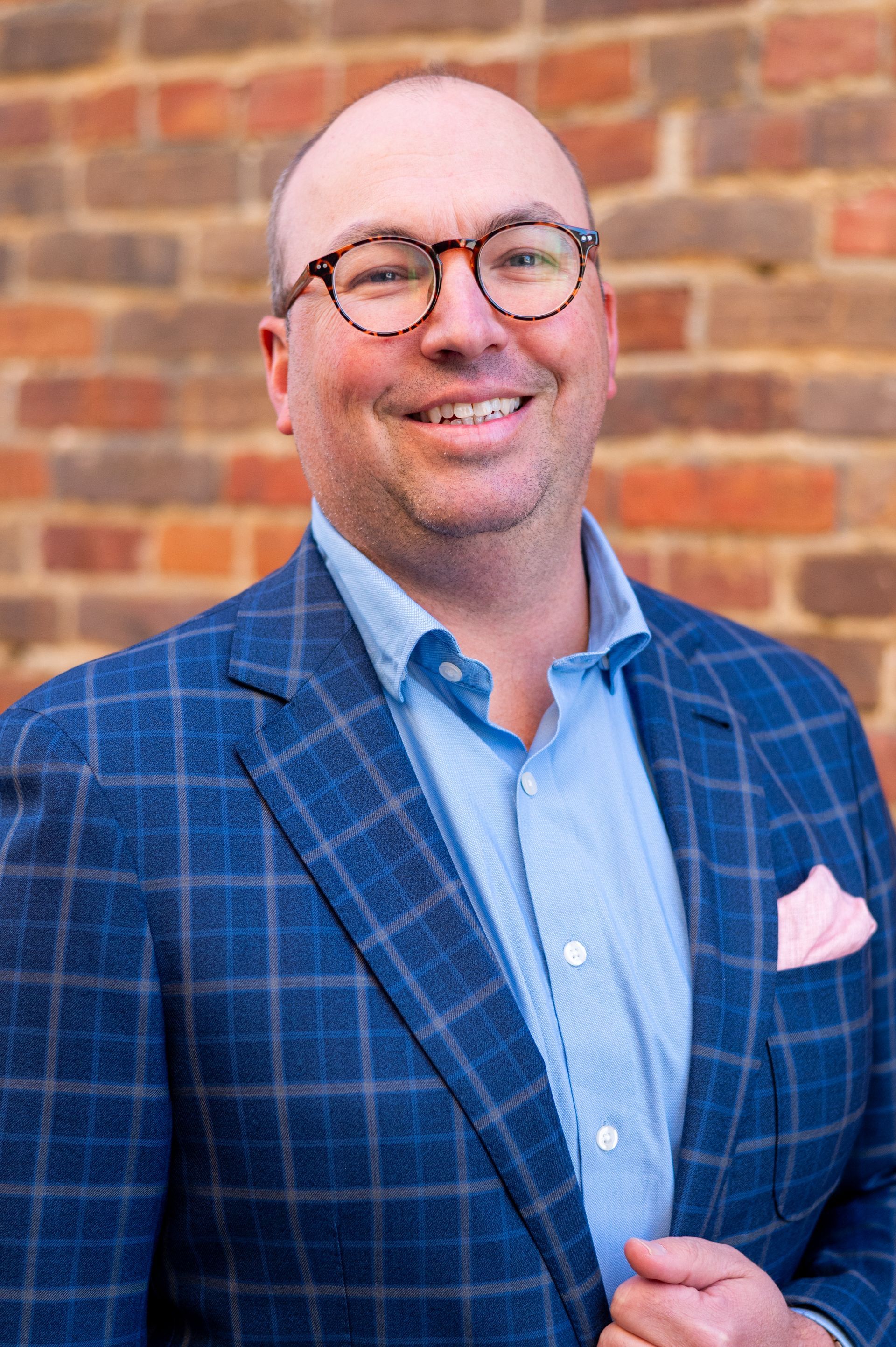 Man in glasses and blue blazer, smiling in front of a brick wall. Pocket square.